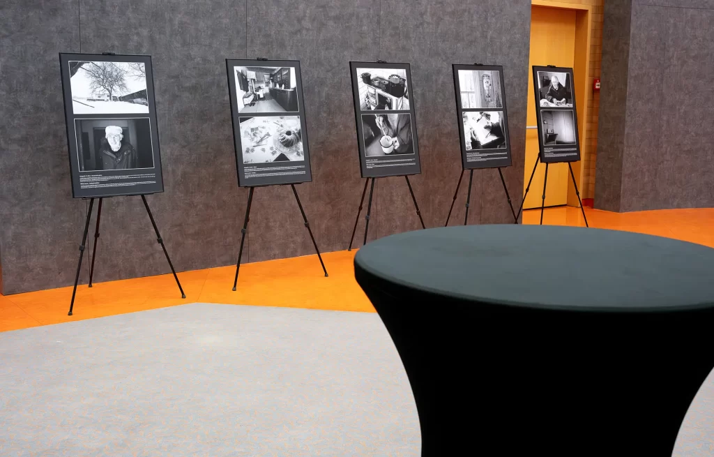 Výstavní sestava s pěti stojany s černobílými fotografiemi umístěnými u texturované šedé zdi připomínající Velkou putovní výstavu. V popředí je kulatý stůl pokrytý černou látkou.