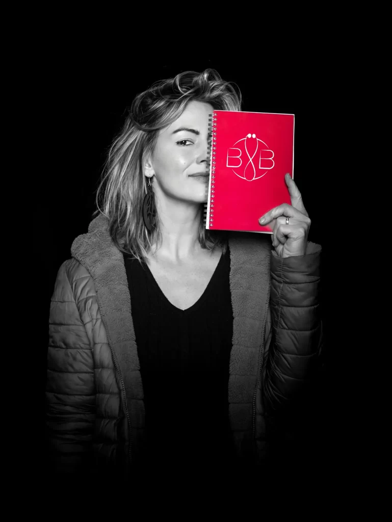 Bára držící červený zápisník s emblémem, který si částečně zakrývá obličej. Má na sobě vycpanou bundu a stojí na černém pozadí.