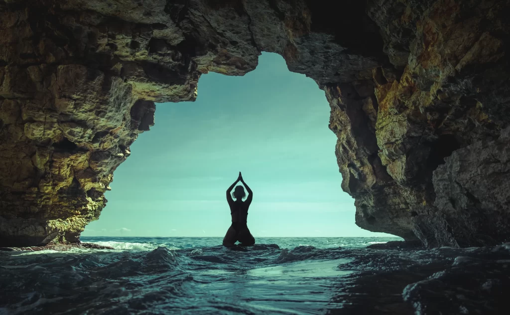 Silueta člověka, který cvičí jógu v jeskyni s výhledem na oceán se světle modrou oblohou viditelnou za skalnatými stěnami.