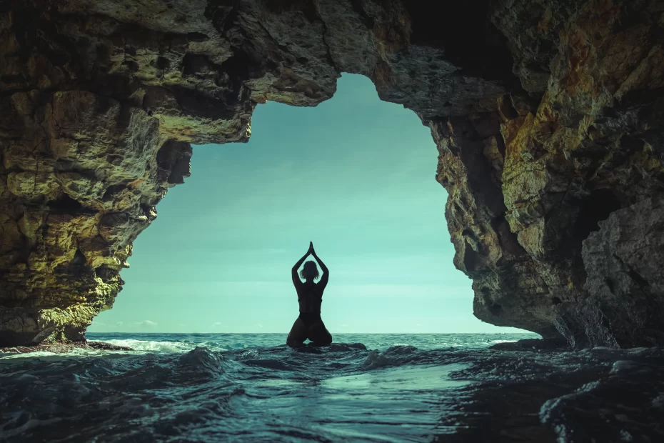 Silueta člověka, který cvičí jógu v jeskyni s výhledem na oceán se světle modrou oblohou viditelnou za skalnatými stěnami.