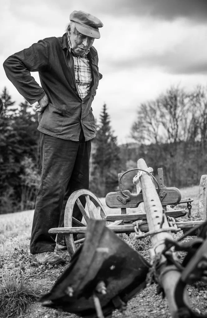 Starší muž v čepici a bundě, stojící venku, shlíží na starý kus zemědělského vybavení. Na pozadí jsou bezlisté stromy a zatažená obloha, připomínající dojemný okamžik zachycený Czech Press Photo 2023.