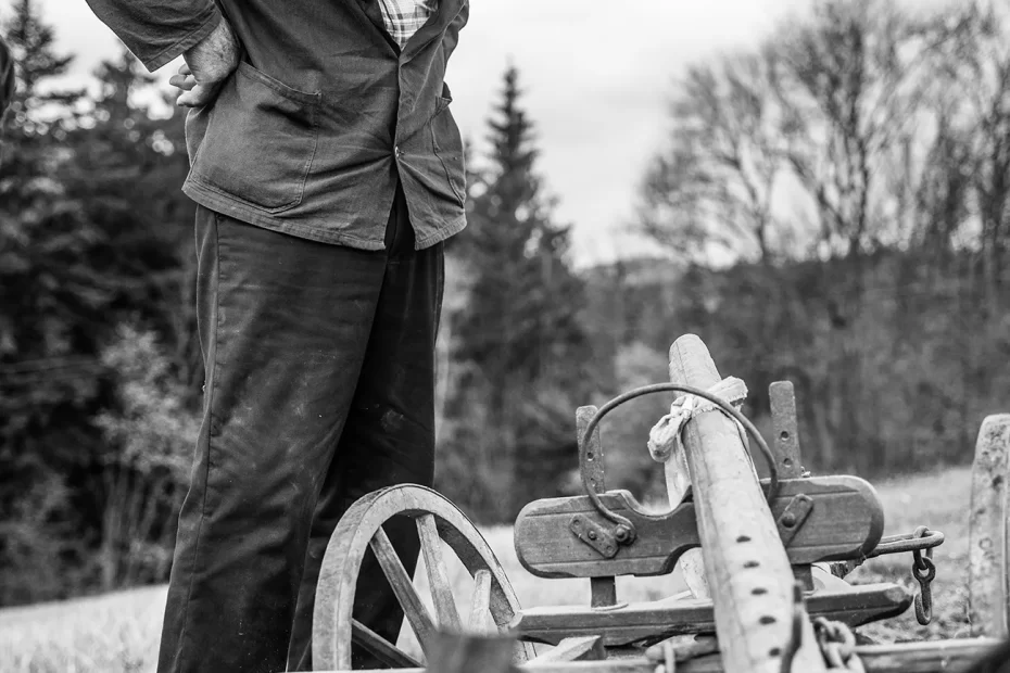 Starší muž v čepici a bundě, stojící venku, shlíží na starý kus zemědělského vybavení. Na pozadí jsou bezlisté stromy a zatažená obloha, připomínající dojemný okamžik zachycený Czech Press Photo 2023.