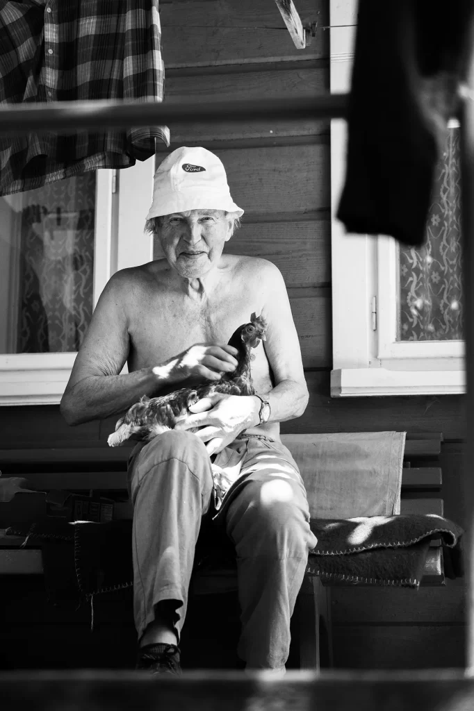 Postarší muž bez košile v klobouku sedí před dřevěnou budovou a jemně drží kuře.