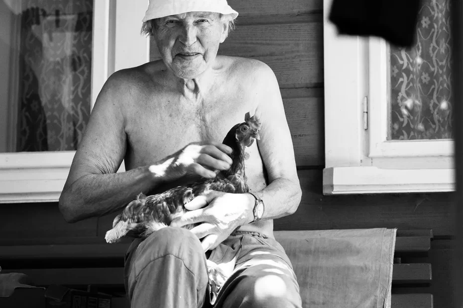 Postarší muž bez košile v klobouku sedí před dřevěnou budovou a jemně drží kuře.